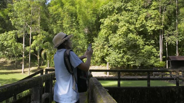 Asian female tourist wearing straw hat and backpack walking on the old concrete bridge over the weir and taking picture with mobile phone. Traveler are enjoy to spent the time in forest park.