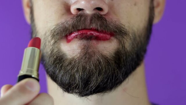 A young guy with a beard paints his lips with red lipstick. Close-up of a bearded man, he painted lips with bright lipstick. A bearded man puts red lipstick on his lips, smiles and smacks his lips.