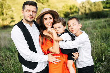 Midshot of a happy family outside on nature. Shot of a loving family. Attractive young man hugs his wife in straw hat while she holds a little boy, the older son is hugging mom and brother