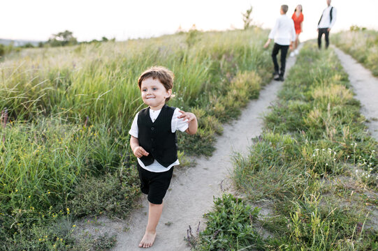 Toddler With A Big Blue Eyes In Suit Vest Runs Outdoors In The Countryside, His Family In Out Of Focus On The Background