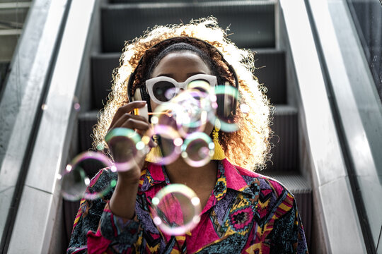 Unrecognizable black woman in headset blowing bubbles sitting on escalator