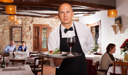 Fototapeta na wymiar Waiter in white shirt holding tray with tasty salad and glass of wine serving for visitors