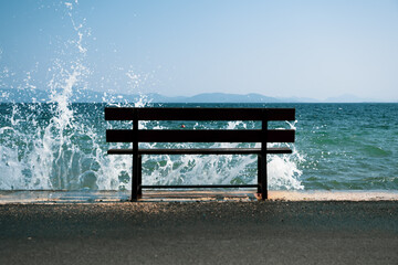 A dark park bench stands at a pier where waves break and spray jumps up in the sun with blue sky and sea.