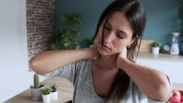 Video of tired young woman suffering neck pain while sitting on the stool in the kitchen at home.