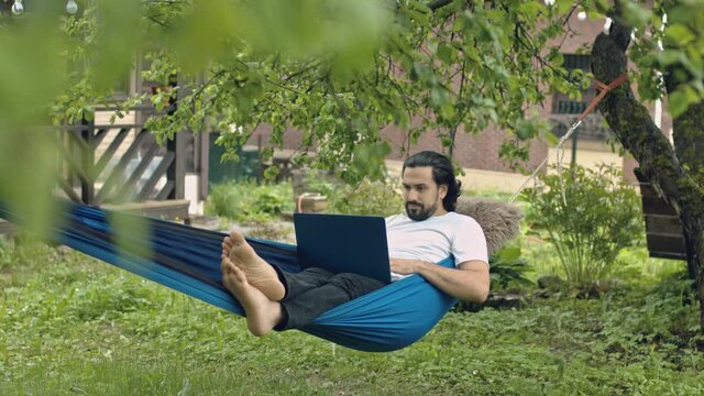 A man works with a laptop in a hammock in a country house. A young guy lies in a hammock with a computer, communicates online, learns remotely under green trees in the country