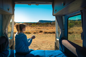 Holidays in Australia. Woman having a coffee in the morning at the back side of a caravan, she wears casual clothes. Motorhome without logos. Vintage look. Kings Canyon, Northern Territory, Australia