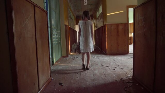 a girl with long black hair and a white dress is walking slowly in a dark corridor. The concept of a horror maniac. Halloween. she is holding a Teddy bear,