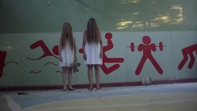 Two girls stand in an abandoned high school gym holding hands, facial hair. Mother and daughter or two sisters. Horror.Abandoned house. Mysticism.