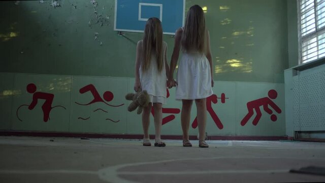 Two girls stand in an abandoned high school gym holding hands, facial hair. Mother and daughter or two sisters. Horror.Abandoned house. Mysticism.