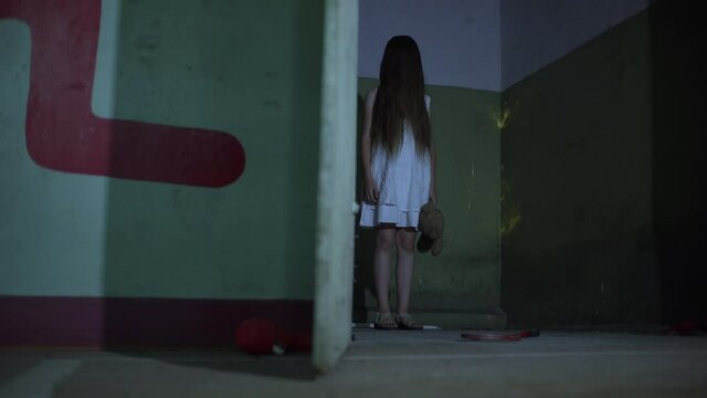 A demonic psycho child in a cursed abandoned house. A little girl dressed in a white dress with a bear toy looks scared at the 4K camera footage