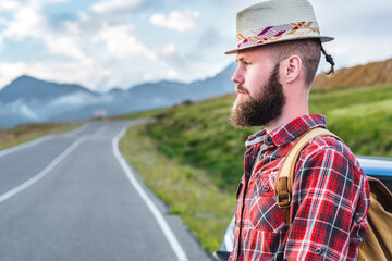 Handsome young man in a straw hat and a plaid shirt, stands on the road, against the backdrop of beautiful mountains. The concept of freedom and hitchhiking