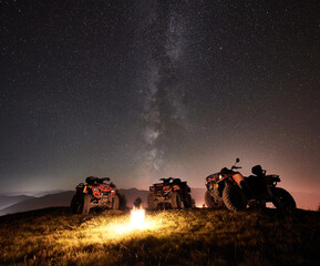 Night picture. Three atv quad motorbikes standing on the top of mountain near burning campfire, under beautiful night sky full of stars and Milky way on background © anatoliy_gleb
