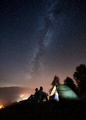 Silhouette of couple travellers having a rest at campfire beside camp and tourist tent under amazing night sky full of stars and Milky way. On the background starry sky, mountains and luminous town