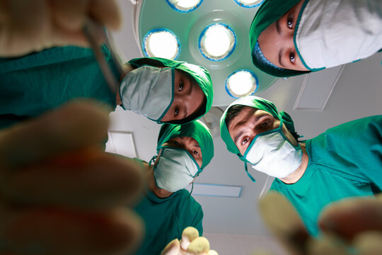 Close Up Four Medical Team In Green Suit Looking Down To Do Operation Above The Patient In Modern Operating Room, The Point Of View From The Patient On Bed.