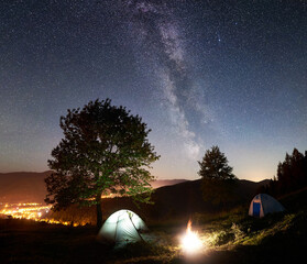Tourist camping near big tree at night. Two tents and bonfire under amazing night sky full of stars and Milky way. On the background incredibly beautiful starry sky, mountains and luminous town