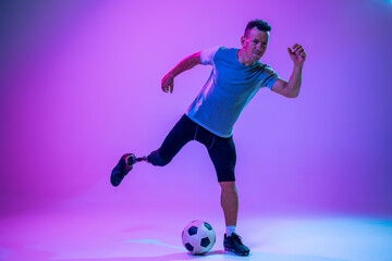 Fototapeta na wymiar Athlete with disabilities or amputee on gradient studio background in neon. Professional male football player with leg prosthesis training in studio. Disabled sport and healthy lifestyle concept.