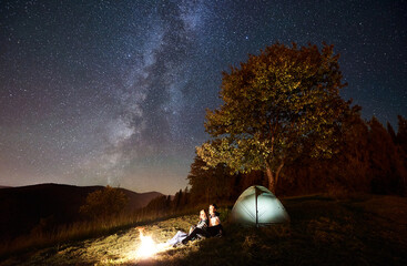 Fototapeta na wymiar Young happy romantic couple hikers resting at bonfire near illuminated tourist tent under amazing night sky full of stars and Milky way. On the background beautiful starry sky, mountains and big tree