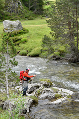 Obraz premium fly fisherman trout fishing with a hiking backpack and an orange jacket in the high mountains in summer