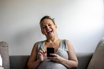 Beautiful pregnant women sitting on sofa with mobile.Young pregnant girl receiving great news on phone.Happy pregnant woman with smartphone in bedroom.Pregnancy, motherhood, technology,people concept