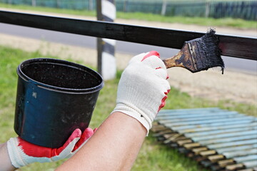 A hand in a white red work glove paints with a brush from paint can a horizontal metal fence impost with black bitumen paint close up in a village on a summer day on old wooden fence background