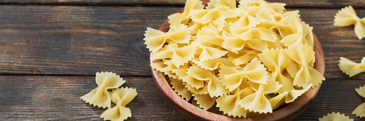 Raw Farfalle macaroni in a wooden bowl on a brown wooden table. Farfalle macaroni close-up. Banner...