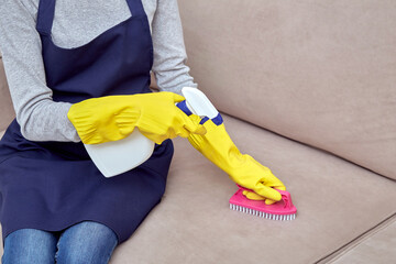 A girl in gloves cleans upholstered furniture with brush and household chemicals