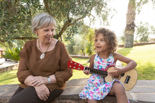 Nipotina Brasiliana suona una chitarra giocattolo vicino alla nonna di origine Europea sedute  in un parco verde