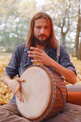 Young hippie man playing on a djembe drum in autumn in the forest.