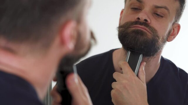 Man shaves his beard using an electric trimmer. Reflection in the mirror