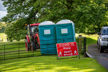 Two plastic portable toilets behind a Covid-19 social distancing warning sign at an outdoor event