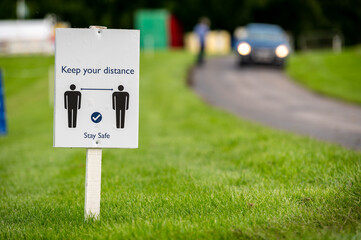 A Covid-19 social distancing sign staked into grass next to a road at an outdoor event