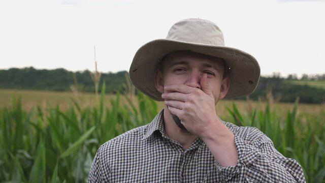 Portrait of tired male farmer yawning against the blurred background of corn field. Exhausted sleepy worker standing in maize meadow and looking into camera. Concept of agricultural business. Close up