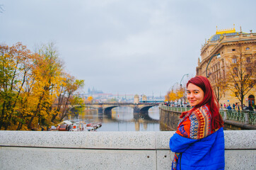 A redhead brazilian woman in her 20s in front of a bridge in Prague, Czechia and smiling to the camera.