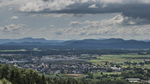Time lapse clouds moving over Ljubljana basin flatland on bright sunny day. Houses and large factory in foreground, hills in background. Zoom in, aerial view