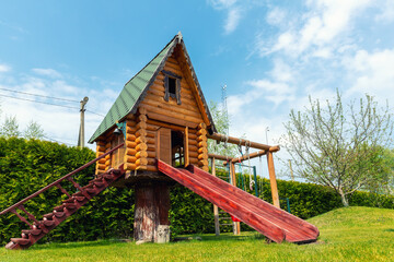 Fototapeta na wymiar Small wood log playhouse hut with stairs ladder and wooden slide on children playground at park or house yard. Green grass lawn garden and blue clear sky in background on bright sunny summer day