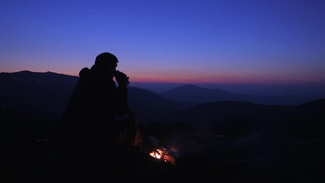 A man sits in the evening by a fire in the mountains and drinks a hot drink from a metal mug. Tourist at the campfire after sunset against the backdrop of beautiful mountains.