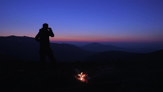 A man drinks a hot drink at night by a campfire in a tourist camp on a mountain pass. A tourist admires the beautiful views of the mountains after sunset. Hiking in the mountains.