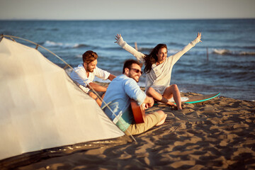 Obraz premium group of caucasian friends singing at the beach, enjoying sunset, smiling, laughing