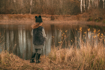 Girl stands near a bush with fallen leaves and looks at the lake