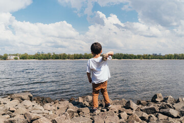 Fototapeta na wymiar back view of cute boy holding rock near blue river