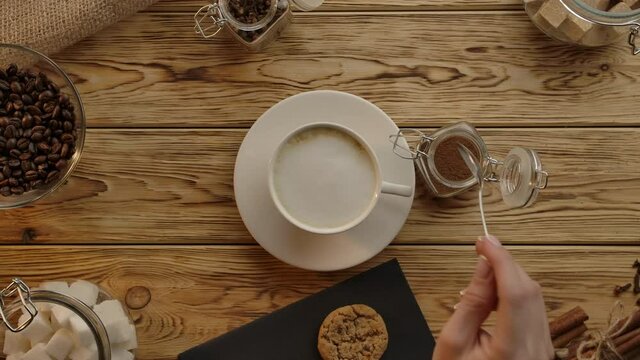 Spices are added to cappuccino: cinnamon or cassia, cardamom, nutmeg powder. Barista hand above coffee cup cooking special fragrant aroma latte with adds on frothed milk surface. Wood desk top view