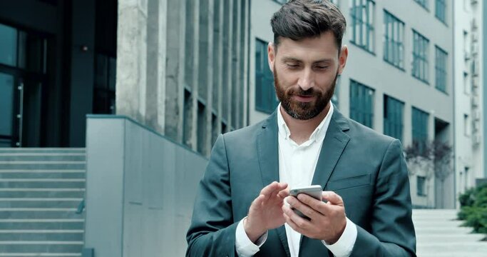 Successful happy Businessman in Luxury grey Suit is texting Messages on his modern Smartphone. Young Man is walking in Business Quartal, having good Mood, looking Satisfied. Successful Lifestyle. Men.
