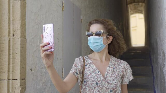 Beautiful curly brunette tourist with a surgical face mask during Covid-19 making a selfie in the pictorestuqe and famous street of Correlo de la rectoria in Sitges, Barcelona. Safe Travel in the new