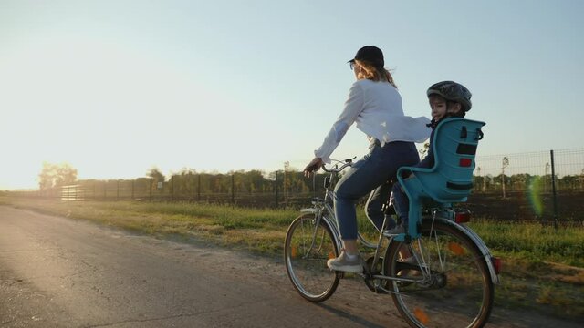 Happy mother with her smiling little son are riding a bike on the road at sunset, slow-motion