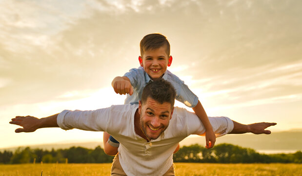 Happy Family Of Father And Child On Field At The Sunset Having Fun Flying On The Shoulder