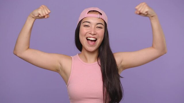 Strong sporty fitness young asian woman girl 20s in casual pink clothes cap posing isolated on pastel purple violet background studio. People lifestyle concept. Look camera showing biceps muscles hand