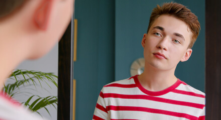 A young teenage guy looks in the mirror at his reflection. Pure beautiful skin and natural beauty of a young man.