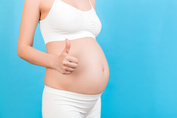 Cropped shot of positive pregnant woman in white underwear showing thumb up cool gesture over her baby bump at blue background. Happy maternity. Copy space