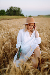 Naklejka na ściany i meble Stylish girl in white dress and hat with book in wheat field. Happy young woman on the sunset or sunrise in summer nature. Fashion and lifestyle concept.