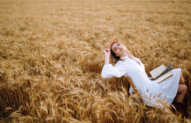 Stylish girl in white dress and hat with book in wheat field. Happy young woman on the sunset or sunrise in summer nature. Fashion and lifestyle concept.